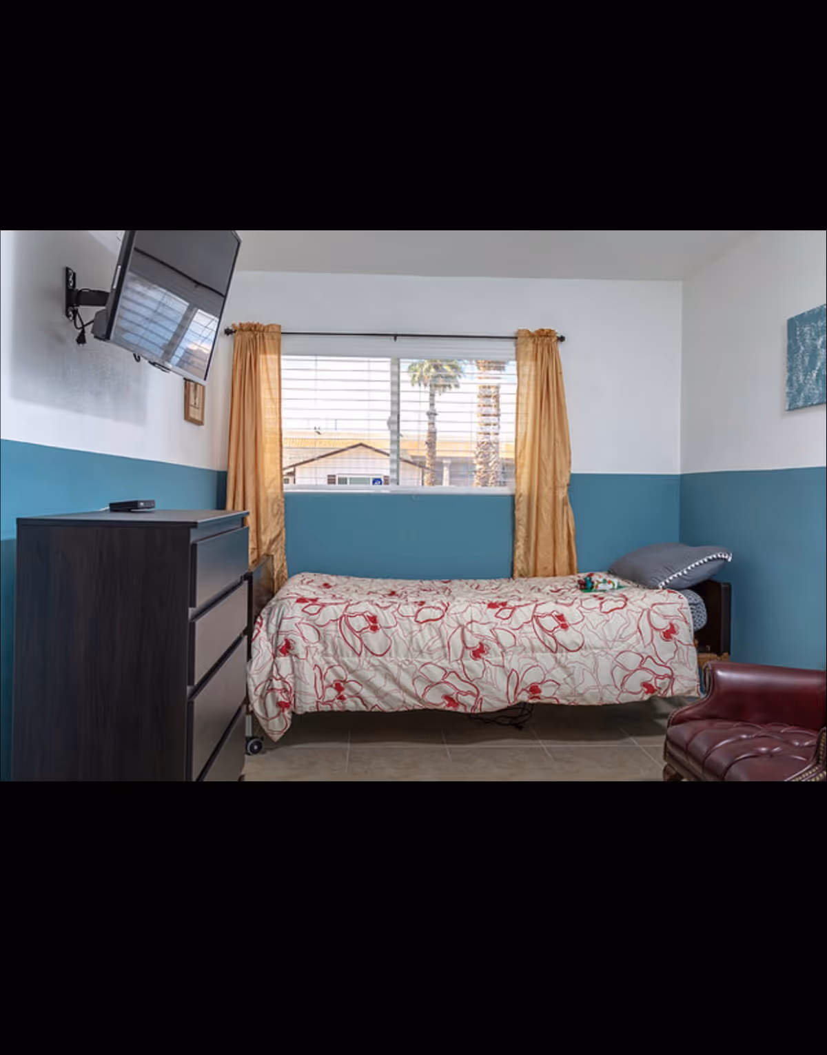 A small bedroom with a single bed covered in a floral patterned bedspread. There is a window with golden curtains behind the bed, showing palm trees and rooftops outside. A dark wooden dresser is on the left side of the bed with a wall-mounted TV above it. A maroon leather chair is partially visible on the right side of the image. The walls are painted white on the upper half and teal on the lower half.