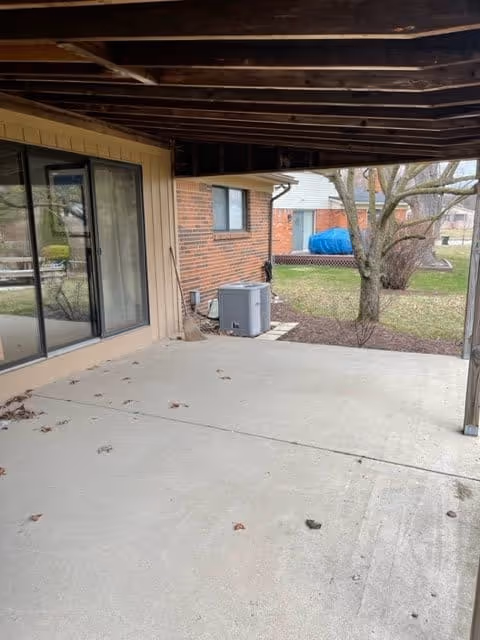 Covered concrete patio area attached to a brick building with a sliding glass door on the left. The patio is empty with some scattered leaves and debris. Beyond the patio, there is a grassy yard with a tree and other buildings visible in the background.