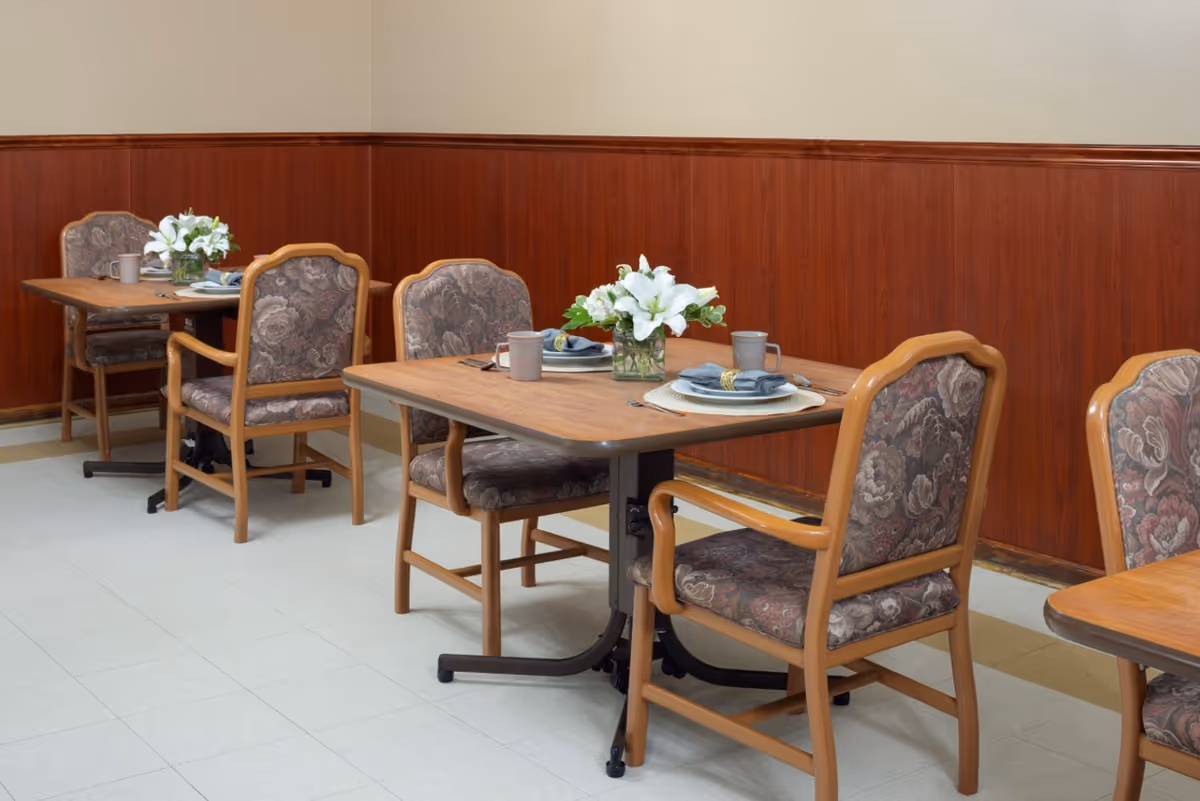 Dining area with wooden tables and padded floral-upholstered chairs, place settings and flower centerpieces against a wood-paneled wall.