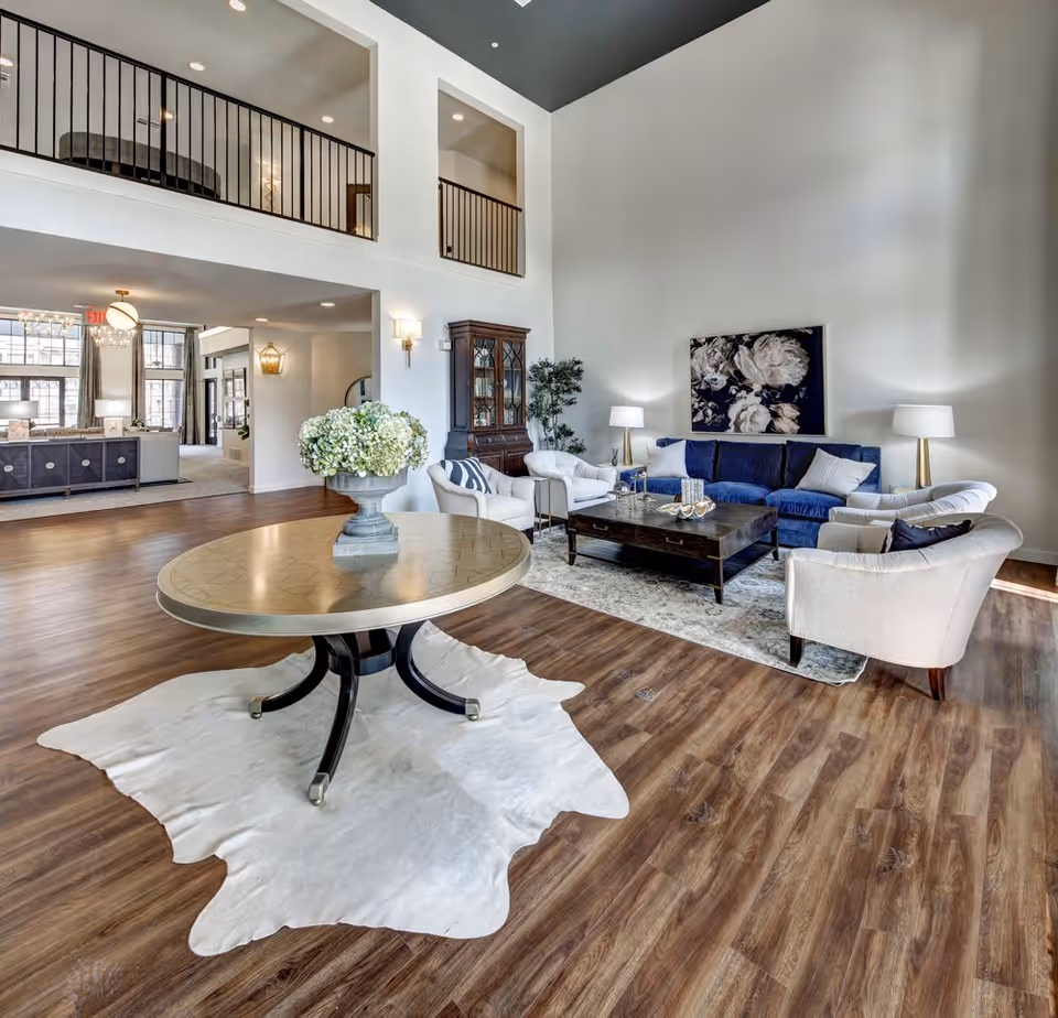 Spacious and well-lit living room area with high ceilings, featuring a blue sofa, four white armchairs arranged around a dark wooden coffee table on a patterned rug. A round table with a flower arrangement sits on a white animal hide rug in the foreground. The room has wooden flooring, a tall cabinet, two floor lamps, and a large floral painting on the wall. An open balcony railing is visible on the upper level, and additional seating and decor can be seen in the background.