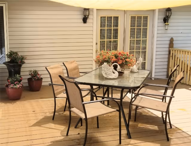 Sunlit outdoor deck patio with a glass-top dining table surrounded by six chairs, potted plants, and swan decorations in front of house doors.