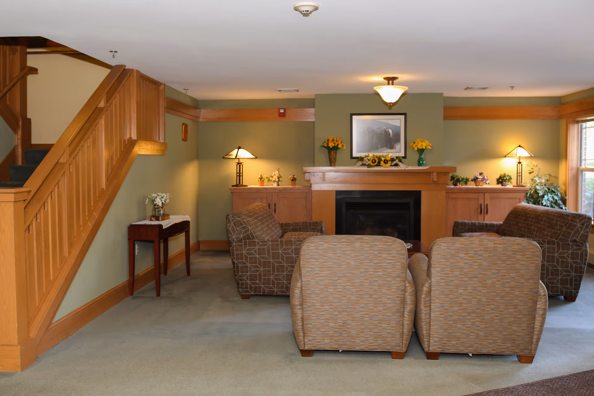 A cozy living room area with four upholstered armchairs arranged around a wooden fireplace. The fireplace mantel is decorated with flower vases and a framed picture above it. There are two wooden cabinets on either side of the fireplace, each with a table lamp and small decorative plants. A staircase with wooden railing is visible on the left side of the room, and a large window on the right lets in natural light.