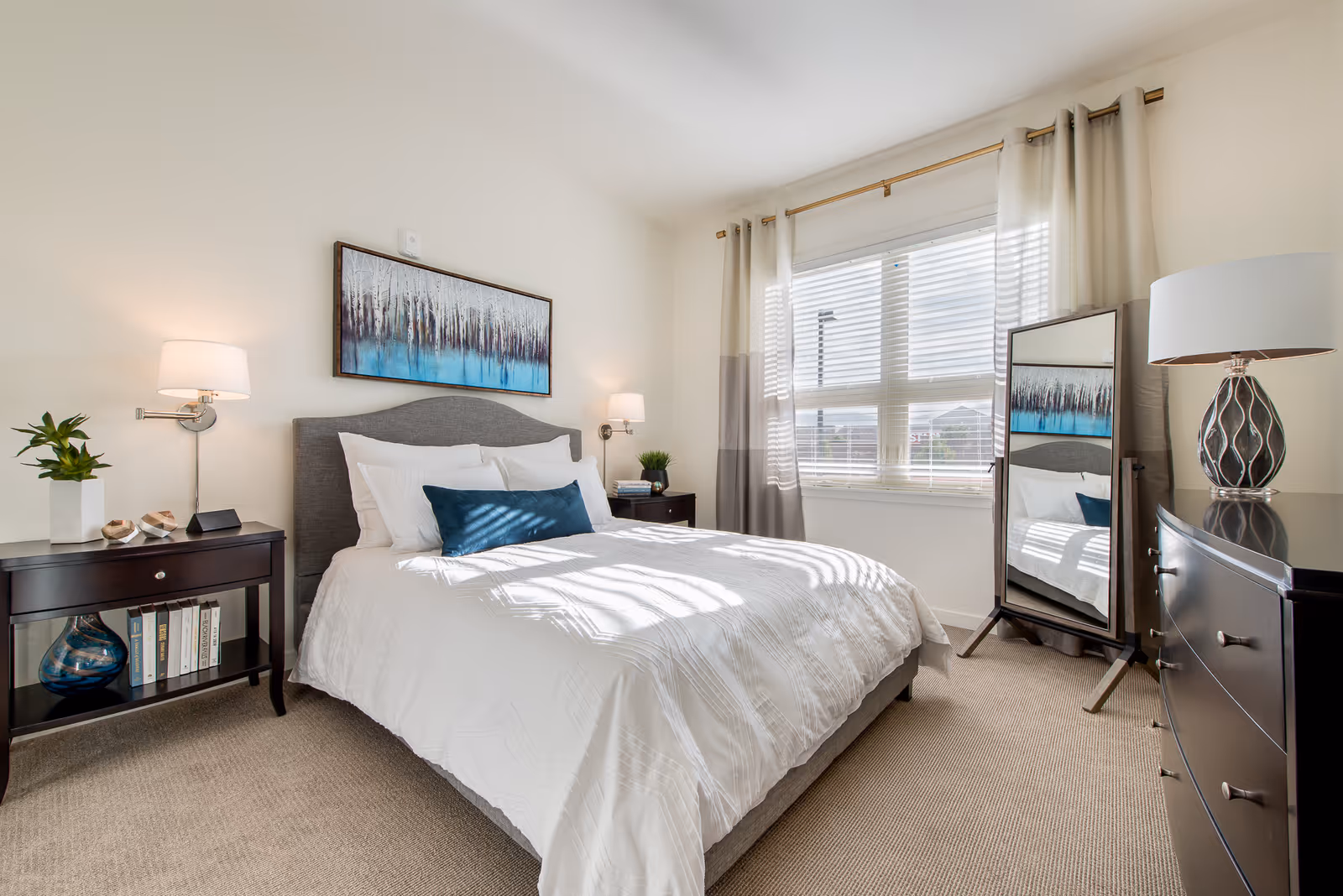 A bright and neatly arranged bedroom with a large window covered by light curtains. The bed has a gray upholstered headboard, white bedding, and a blue accent pillow. On either side of the bed are dark wooden nightstands with lamps, books, and small plants. A full-length mirror stands near the window, reflecting part of the room. The walls are light-colored, and a framed artwork featuring trees and a blue gradient hangs above the bed.