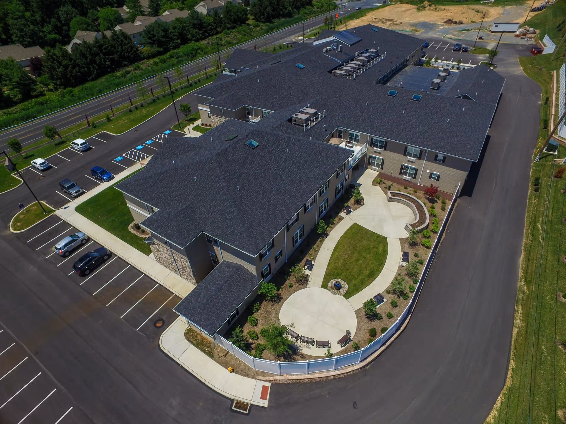 Aerial view of Heather Glen At Ardenwoods senior living facility showing a large two-story building with a dark roof, surrounding parking lot with several cars, landscaped garden area with pathways, benches, and a circular paved section enclosed by a white fence.