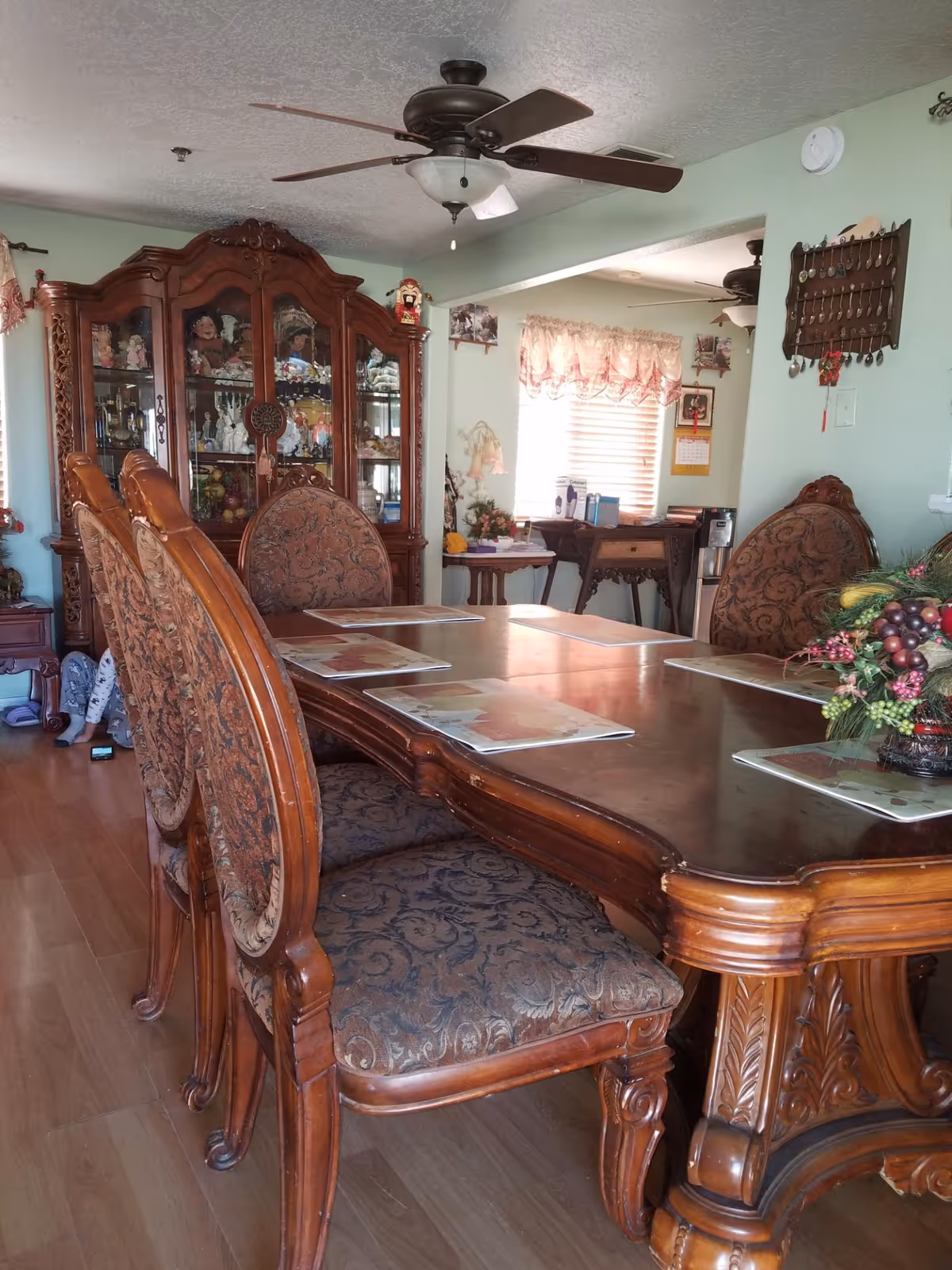 Ornate wooden dining table with upholstered chairs, placemats, a floral centerpiece, china cabinet and ceiling fan in a home dining room.