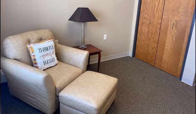 A beige armchair with a matching ottoman in a carpeted room. The armchair has a decorative pillow with the words 'THANKFUL grateful BLESSED'. Next to the chair is a wooden side table with a lamp that has a dark lampshade. Behind the chair is a wall with an electrical outlet, and to the right are wooden double doors.