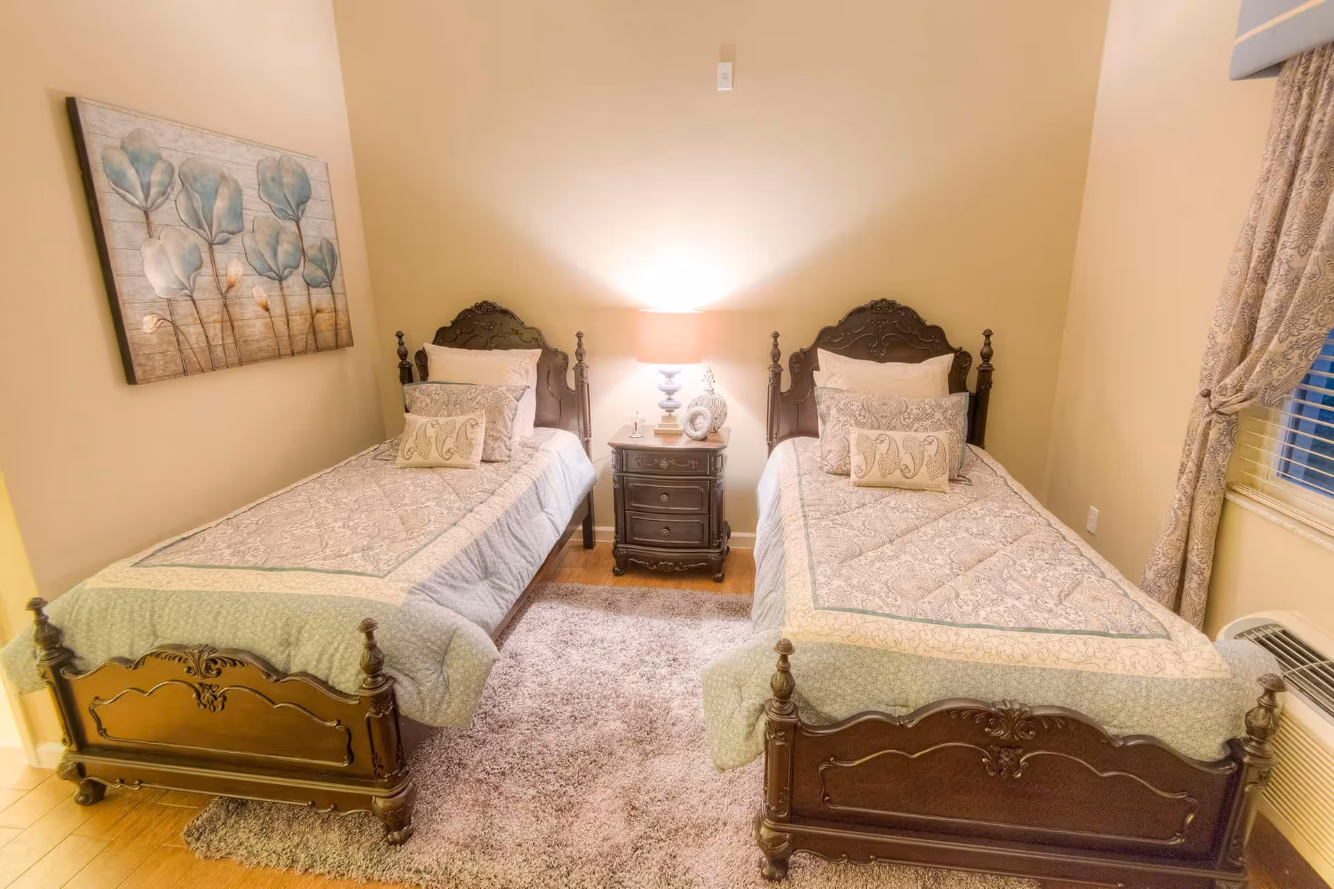 A bedroom with two single beds featuring ornate dark wooden headboards and footboards. Each bed is made with patterned bedspreads and multiple pillows. A small dark wooden nightstand with three drawers is placed between the beds, holding a lit table lamp, a small clock, and decorative items. A light-colored rug is on the wooden floor beneath the beds. On the left wall, there is a painting of blue and beige flowers, and on the right side, a window with patterned curtains and an air conditioning unit below it.