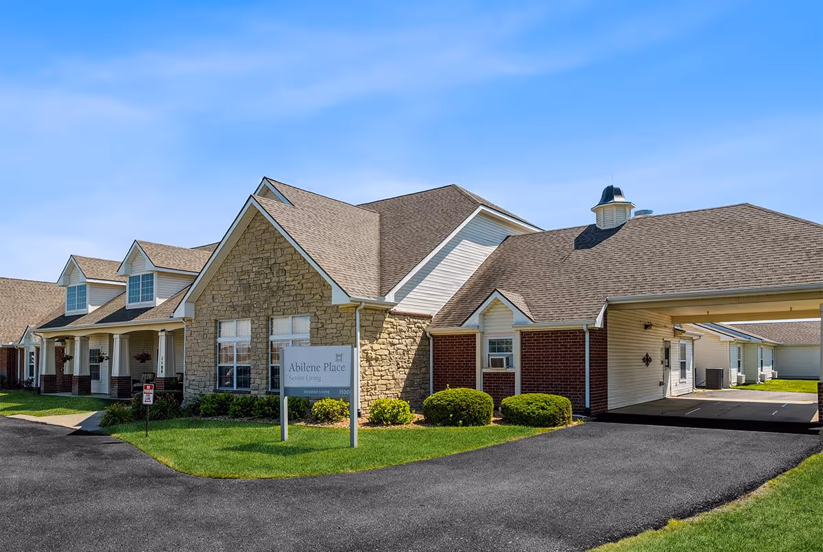 Exterior view of Abilene Place South senior living facility showing a single-story building with stone and brick facade, multiple windows, a covered driveway, and well-maintained green lawn under a clear blue sky.