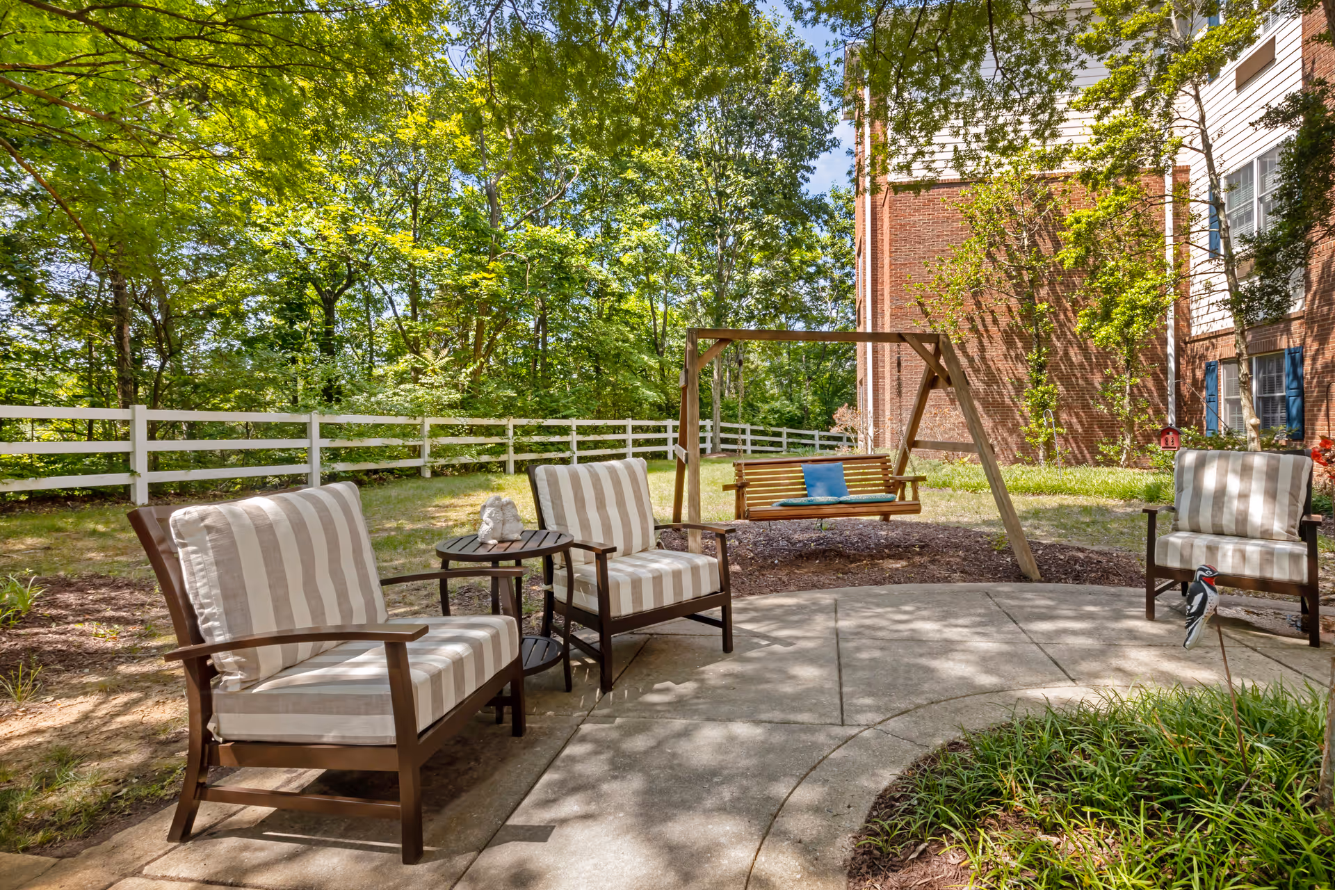 Outdoor seating area with three cushioned chairs arranged around a small round table on a curved concrete patio. Behind the chairs is a wooden swing with a blue cushion, set in a garden area with green trees, grass, and a white fence. Part of a brick and white building is visible on the right side.