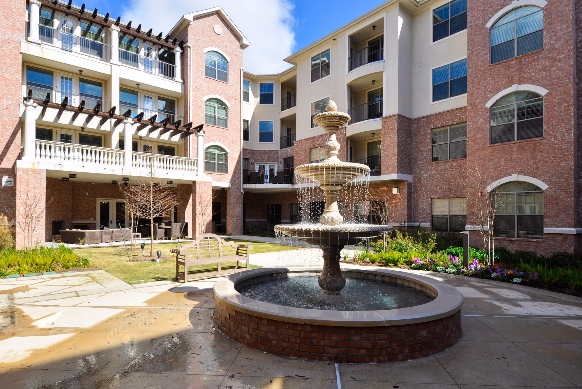 Outdoor courtyard area of The Abbey at Westminster Plaza featuring a multi-tiered water fountain in the center, surrounded by a circular brick base. There is a wooden bench nearby, landscaped flower beds, and a multi-story building with balconies and large windows in the background under a partly cloudy sky.