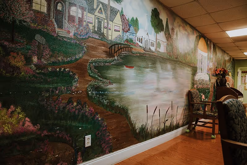 A hallway in an assisted living facility featuring a large mural on the wall depicting a scenic village with houses, a pond, a small bridge, and trees. The floor is wooden, and there are chairs with floral upholstery along the wall.