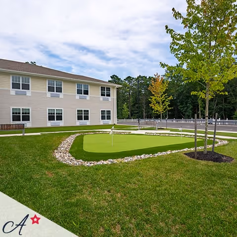 Outdoor view of a two-story assisted living facility building with multiple windows. In the foreground, there is a small putting green surrounded by rocks and grass, with a few young trees planted nearby. The sky is partly cloudy.