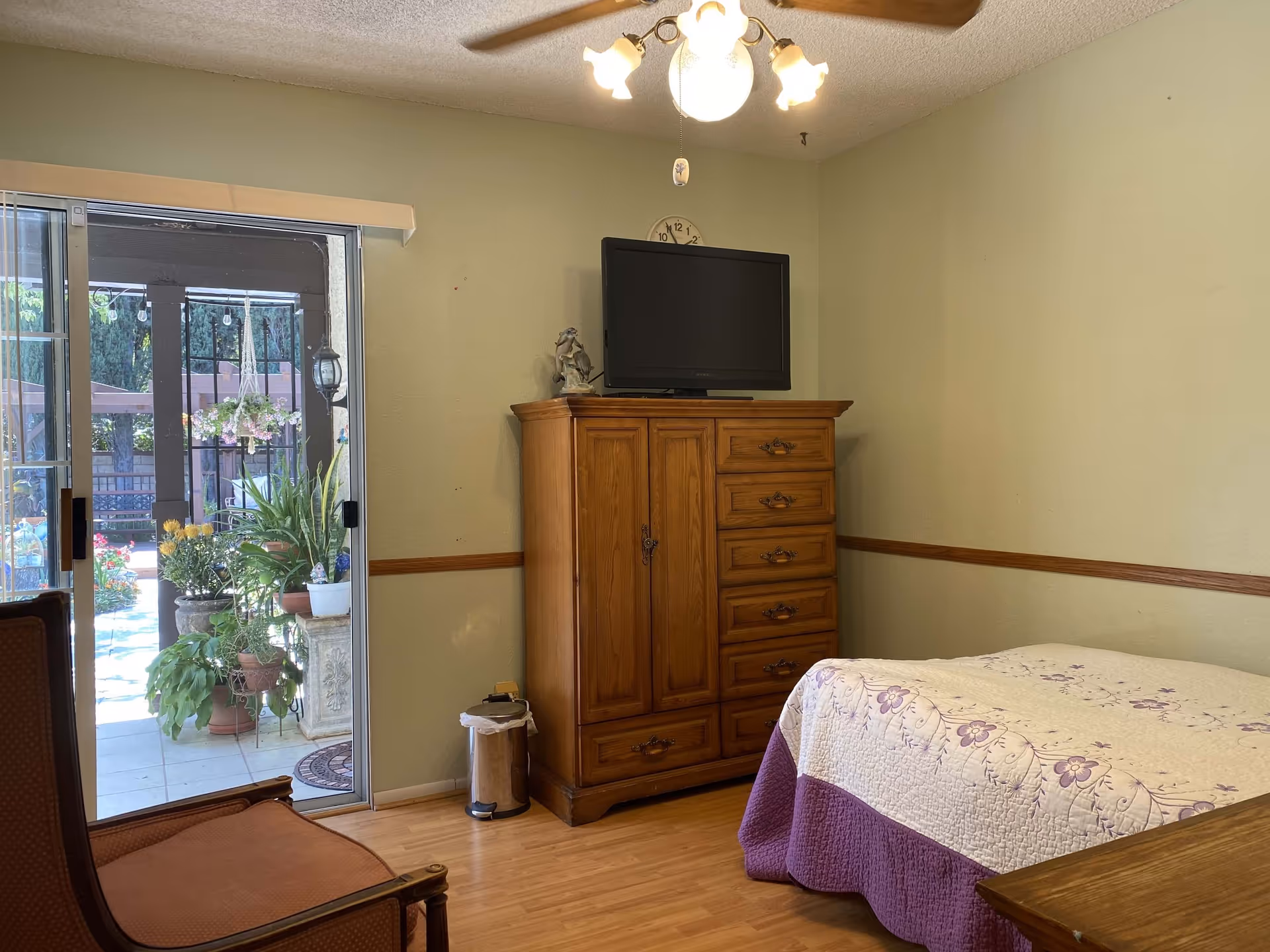 A bedroom with a wooden dresser topped with a flat-screen TV and a small figurine. The bed has a white and purple floral quilt. There is a wooden chair with a brown cushion and a sliding glass door leading to an outdoor patio with various potted plants and hanging flowers. The room has light green walls, a ceiling fan with lights, and wooden flooring.
