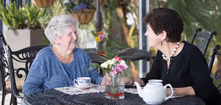 Two women sit at an outdoor patio table with a teapot, teacup, and a small vase of flowers, conversing.