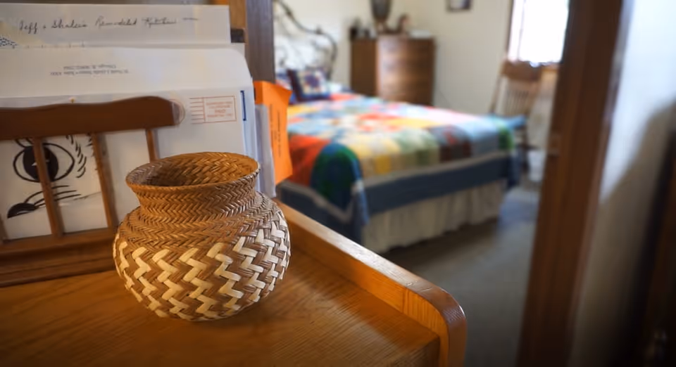 Woven wicker basket on a wooden dresser in the foreground with a blurred bedroom and colorful quilted bed visible in the background.