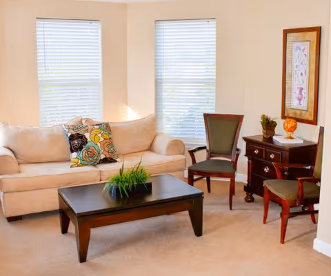 A bright living room with a beige sofa adorned with colorful floral pillows, two wooden chairs with green cushions, a dark wooden coffee table with a small green plant, and a matching dark wooden side table with decorative items and a framed artwork on the wall. Two windows with white blinds allow natural light into the room.