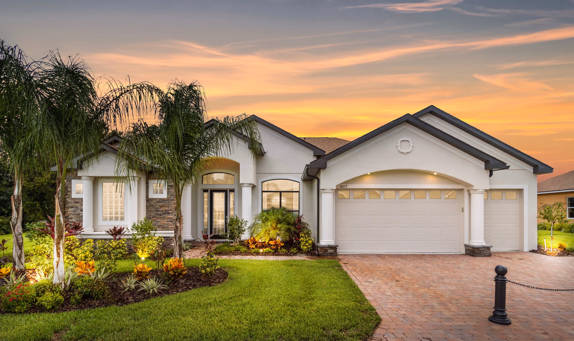 Front exterior of a single-story suburban house with a three-car garage, landscaped yard and palm trees at sunset.