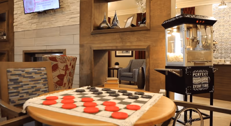 Cozy common area with a checkers game set up on a round wooden table, surrounded by patterned chairs. In the background, there is a popcorn machine with the text 'Perfect Popcorn Every Time' and a fireplace with a mounted TV above it. The room has warm wooden accents and comfortable seating.