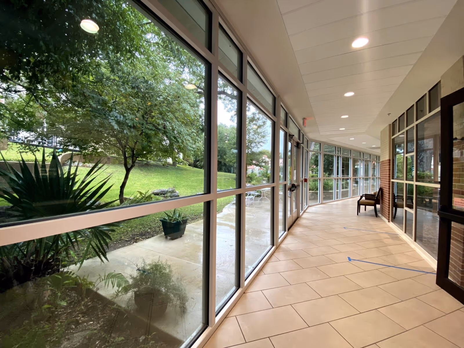 A bright indoor hallway with large floor-to-ceiling windows on the left side showing a green outdoor garden area with trees and plants. The hallway has beige tiled flooring, recessed ceiling lights, and a single chair placed near the windows on the right side.