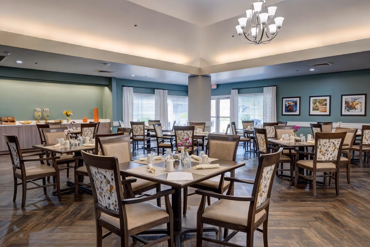 A spacious dining room with multiple wooden tables and chairs arranged neatly. Each table is set with napkins, cups, and small flower vases. The room has large windows with white curtains allowing natural light to enter. The walls are painted green and decorated with framed pictures. A chandelier hangs from the ceiling, and there is a buffet area with beverage dispensers and decorative items in the background.