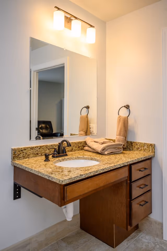 A bathroom vanity with a granite countertop, an undermount sink, and a dark faucet. Above the sink is a large rectangular mirror with three light fixtures mounted above it. A beige towel is hanging on a towel ring on the wall, and folded beige towels are placed on the countertop. The vanity has wooden drawers and a partial wooden panel underneath. The floor is tiled and a doorway is visible in the mirror reflection.