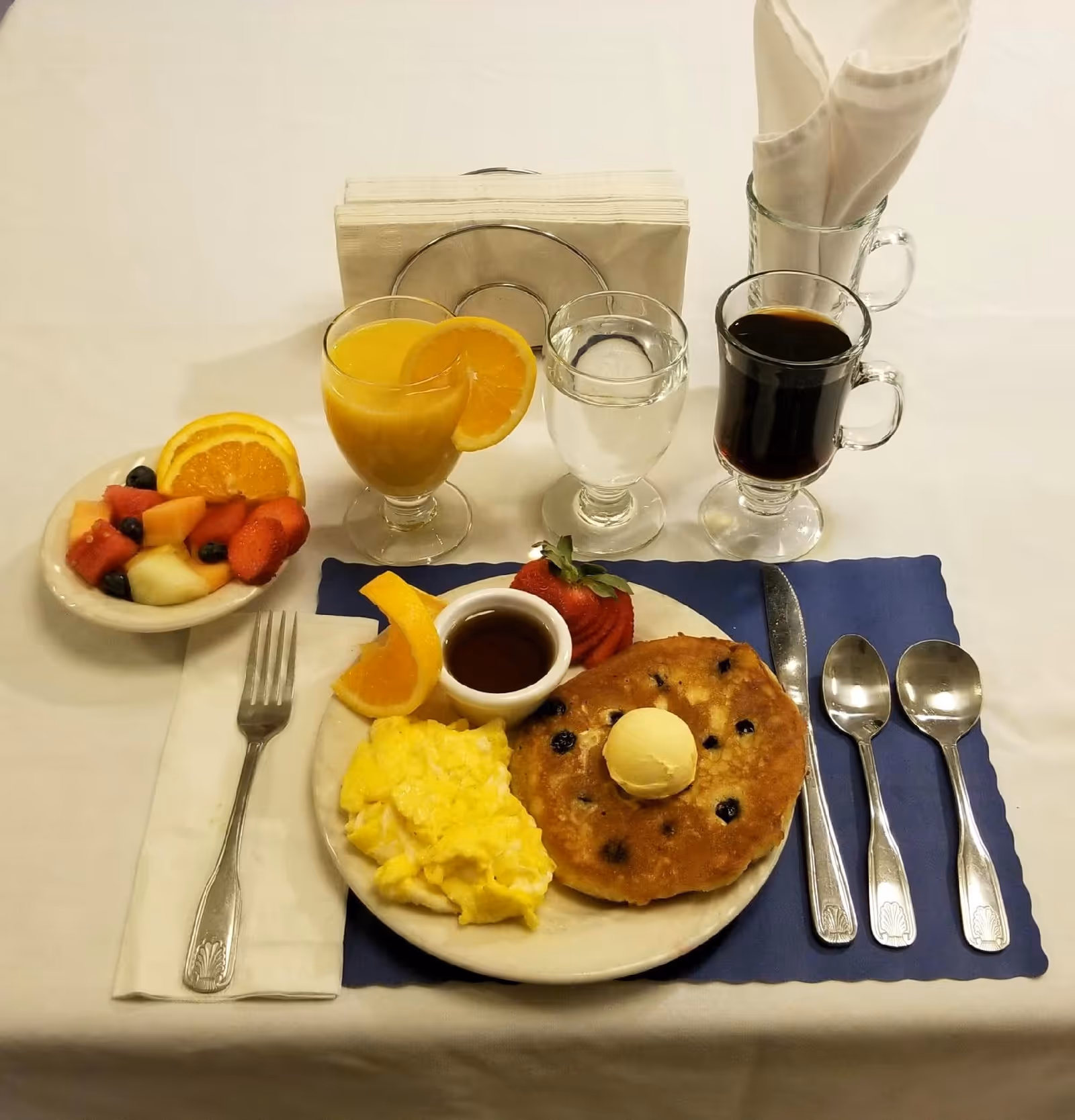 A breakfast meal set on a white tablecloth with a plate containing scrambled eggs, a blueberry pancake topped with a scoop of butter, a strawberry, an orange slice, and a small cup of syrup. Next to the plate is a fork on a napkin, and on the right side are a knife and two spoons on a blue placemat. Behind the plate are three glasses containing orange juice with an orange slice, water, and black coffee. A small bowl of mixed fruit with orange slices, strawberries, cantaloupe, and blueberries is also present. Napkins are held in a glass container in the background.