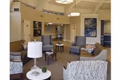 A cozy nursing home common area with several upholstered chairs and sofas arranged around small tables. The room features neutral tones, carpeted floors, modern ceiling lights, and framed artwork on the walls. A small lamp and decorative plant are on a side table in the foreground.