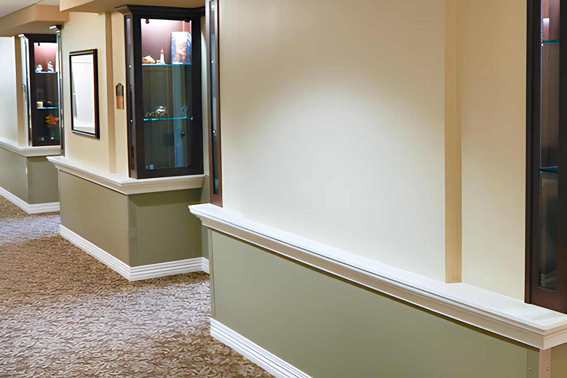 A well-lit hallway in a senior living facility with beige and green walls, carpeted floor, and built-in glass display cabinets showcasing decorative items.