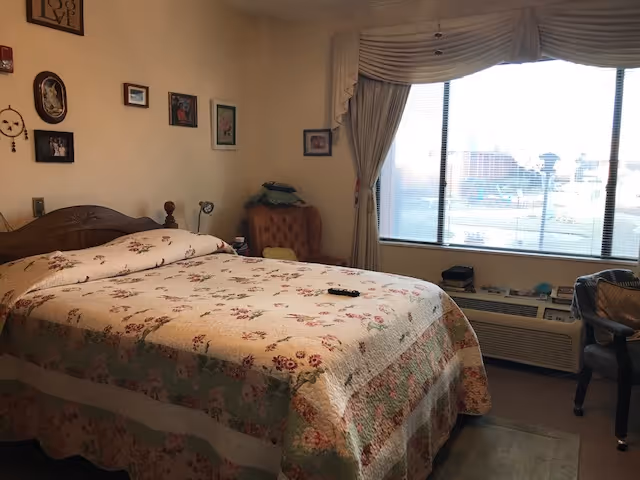 A bedroom with a large bed covered in a floral quilt. There is a wooden headboard, a cushioned armchair in the corner, and several framed pictures hanging on the wall. A large window with curtains lets in natural light, and there is a heating or air conditioning unit beneath the window. A chair is positioned near the window.