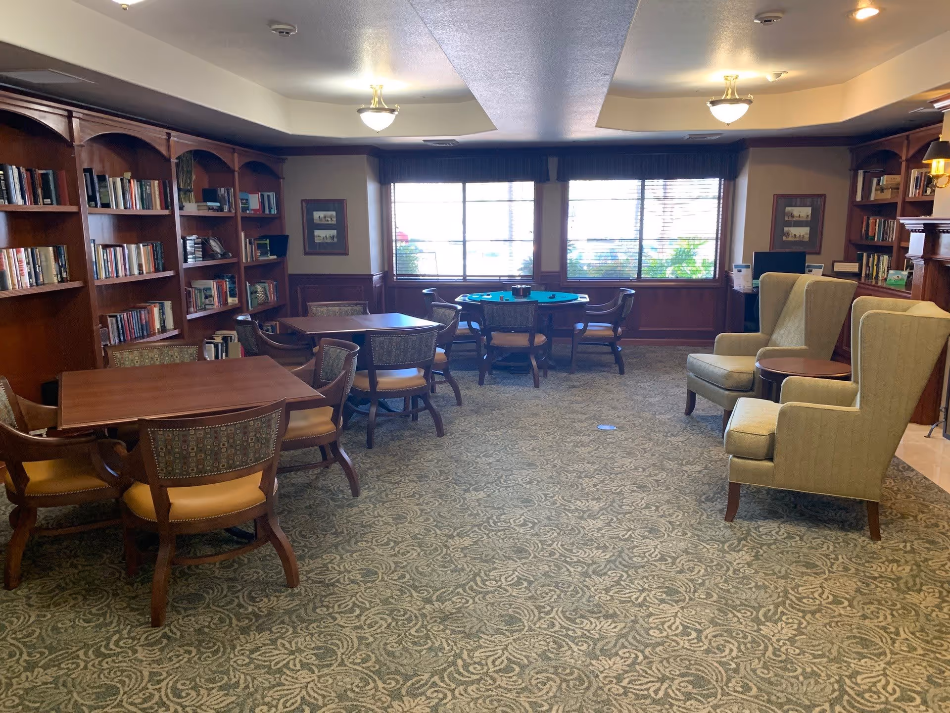 A cozy common area with wooden bookshelves filled with books along the left wall, several wooden tables with chairs, two upholstered armchairs with a small round table between them, and large windows letting in natural light at the far end of the room.