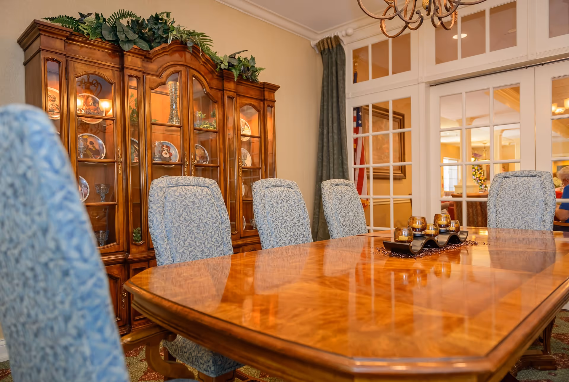 A polished wooden dining table with six upholstered chairs featuring a blue floral pattern. On the table is a decorative centerpiece with candles. Behind the table is a wooden china cabinet displaying plates and glassware. In the background, there are glass-paneled double doors and a glimpse of another room with an American flag and framed artwork.