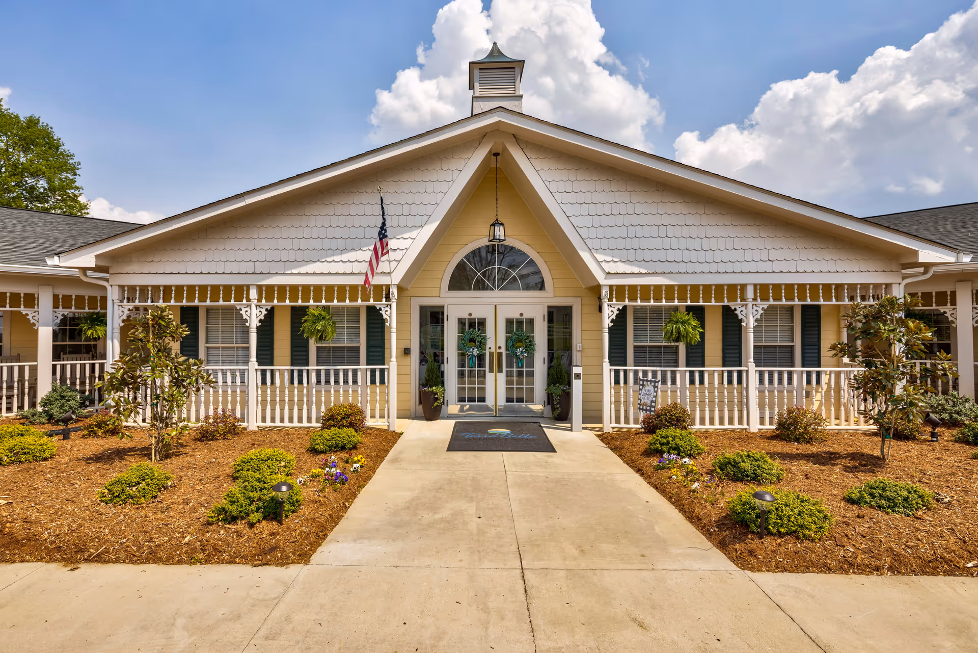 Front exterior view of TerraBella Asheboro facility with a wide concrete walkway leading to double glass doors adorned with wreaths. The building features a peaked roof with decorative shingles, white railings, hanging plants, and an American flag. The landscaped area includes small bushes, flowers, and mulch under a partly cloudy sky.