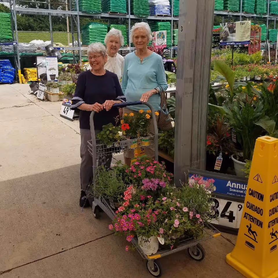 Three elderly women standing outdoors at a garden center with a cart full of colorful flowering plants. They are smiling and appear to be enjoying their time shopping for plants. Behind them are shelves stacked with bags of soil and gardening supplies.