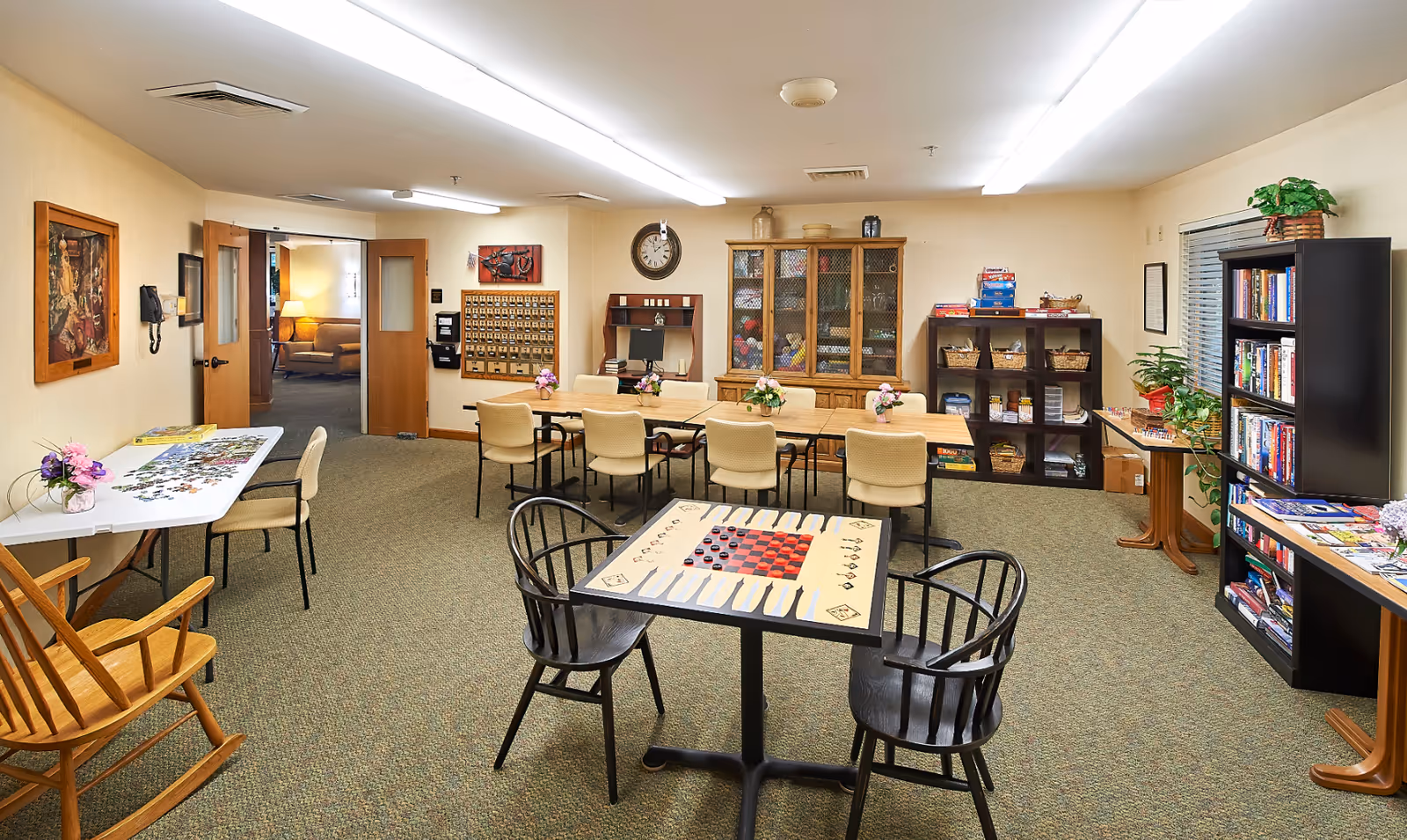 A bright communal activity room with multiple tables and chairs, bookshelves, and game tables in a senior living facility.
