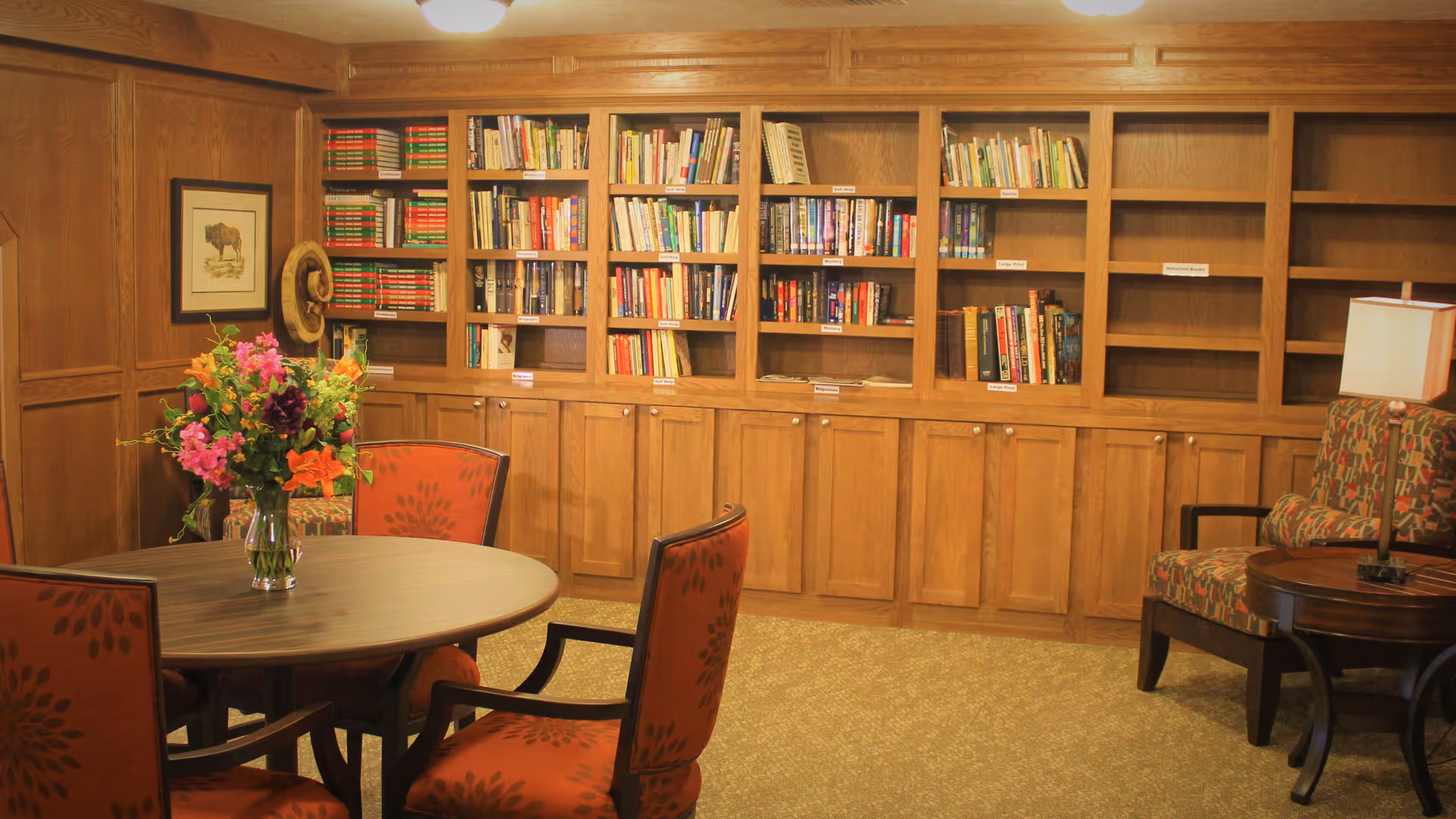 A cozy library room with wooden bookshelves filled with books, a round wooden table with a vase of colorful flowers, and several upholstered chairs with patterned fabric. There is a framed picture on the wall and a table lamp on a side table next to one of the chairs.