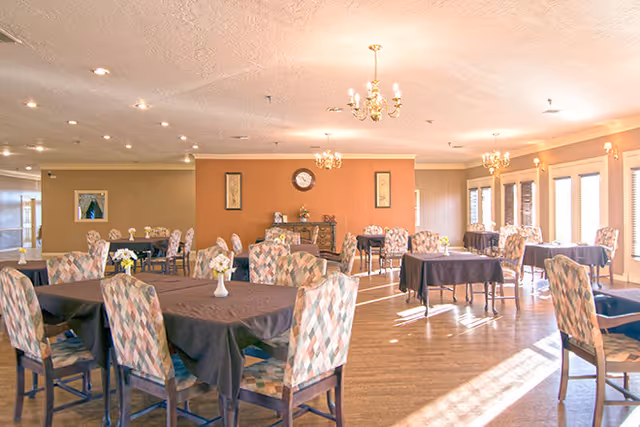 A spacious dining room with multiple tables covered in dark tablecloths and surrounded by patterned upholstered chairs. The room has wooden flooring, chandeliers hanging from the ceiling, and large windows allowing natural light to fill the space. There is a clock and framed artwork on the walls.