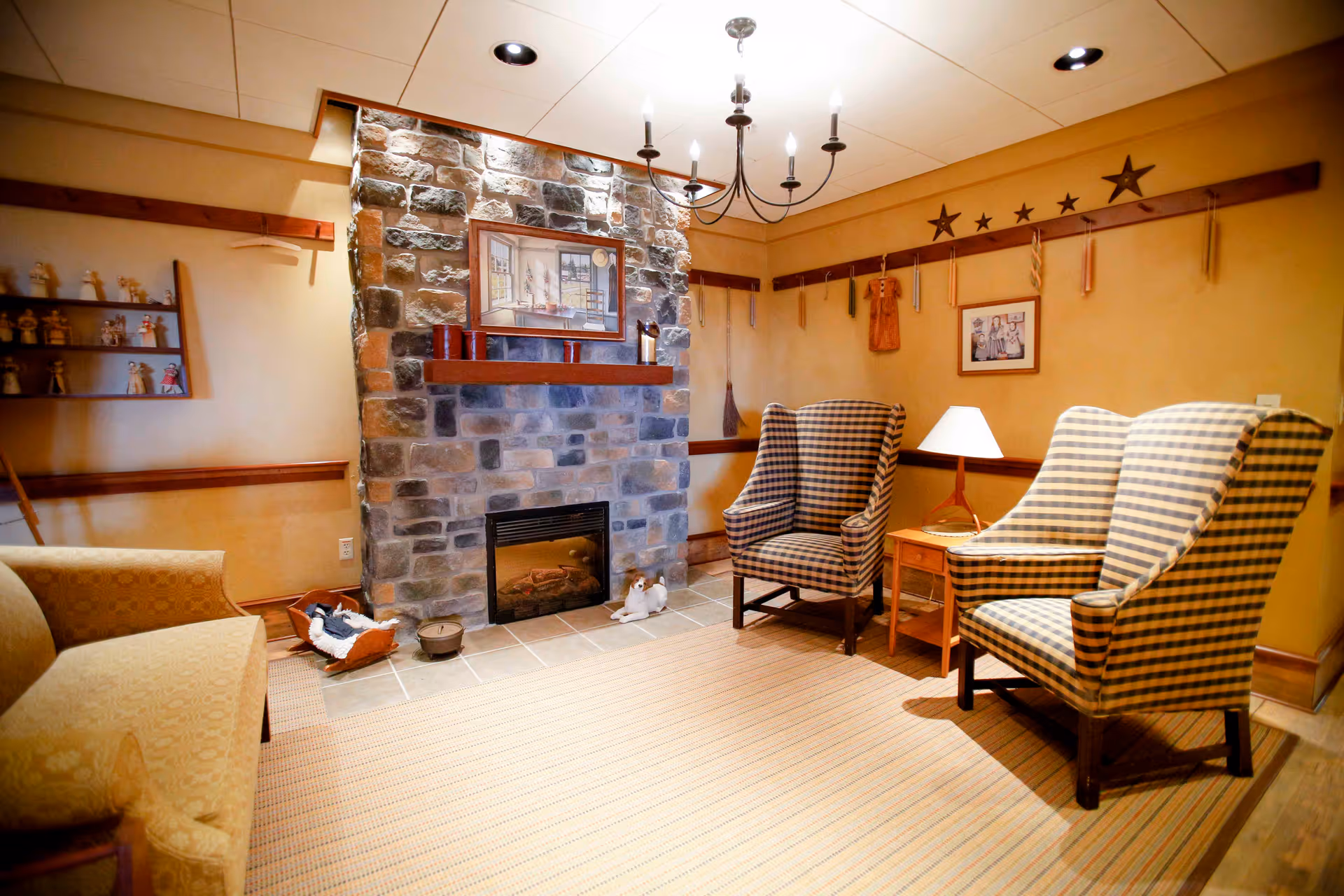 A cozy living room with a stone fireplace in the center, flanked by two checkered armchairs on the right and a beige sofa on the left. The room has warm yellow walls decorated with wooden stars, framed pictures, and small shelves holding figurines. A small wooden side table with a lamp is placed between the armchairs.