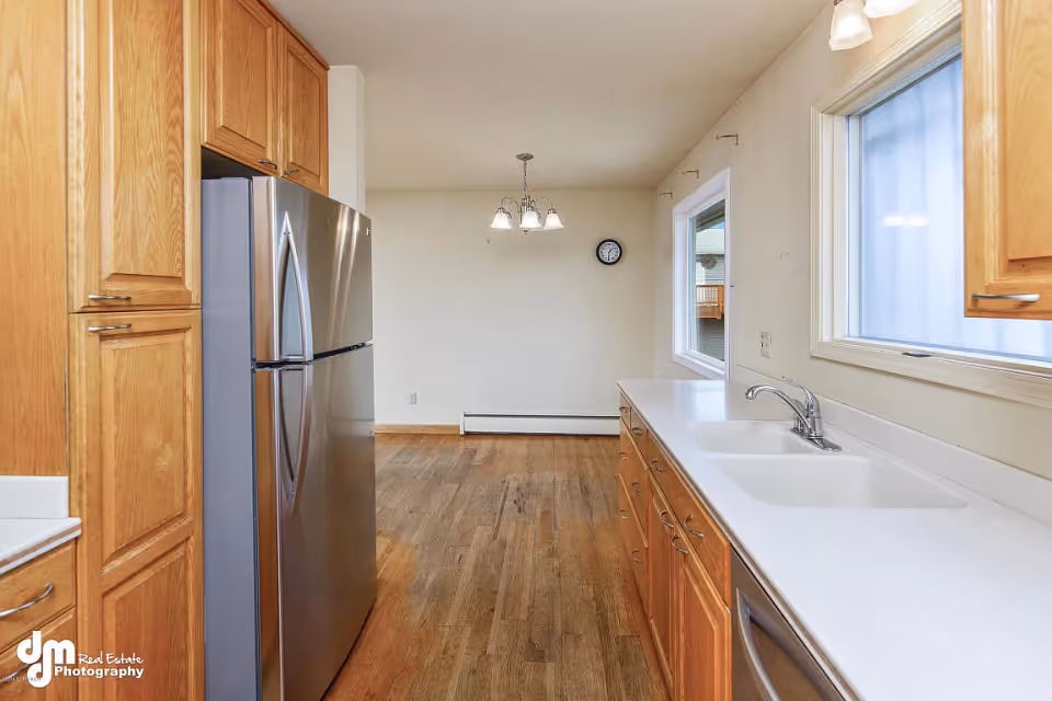 A kitchen interior featuring wooden cabinets, a stainless steel refrigerator, a white countertop with a double sink, and two windows. The kitchen opens into a dining area with a hanging light fixture and a wall clock.