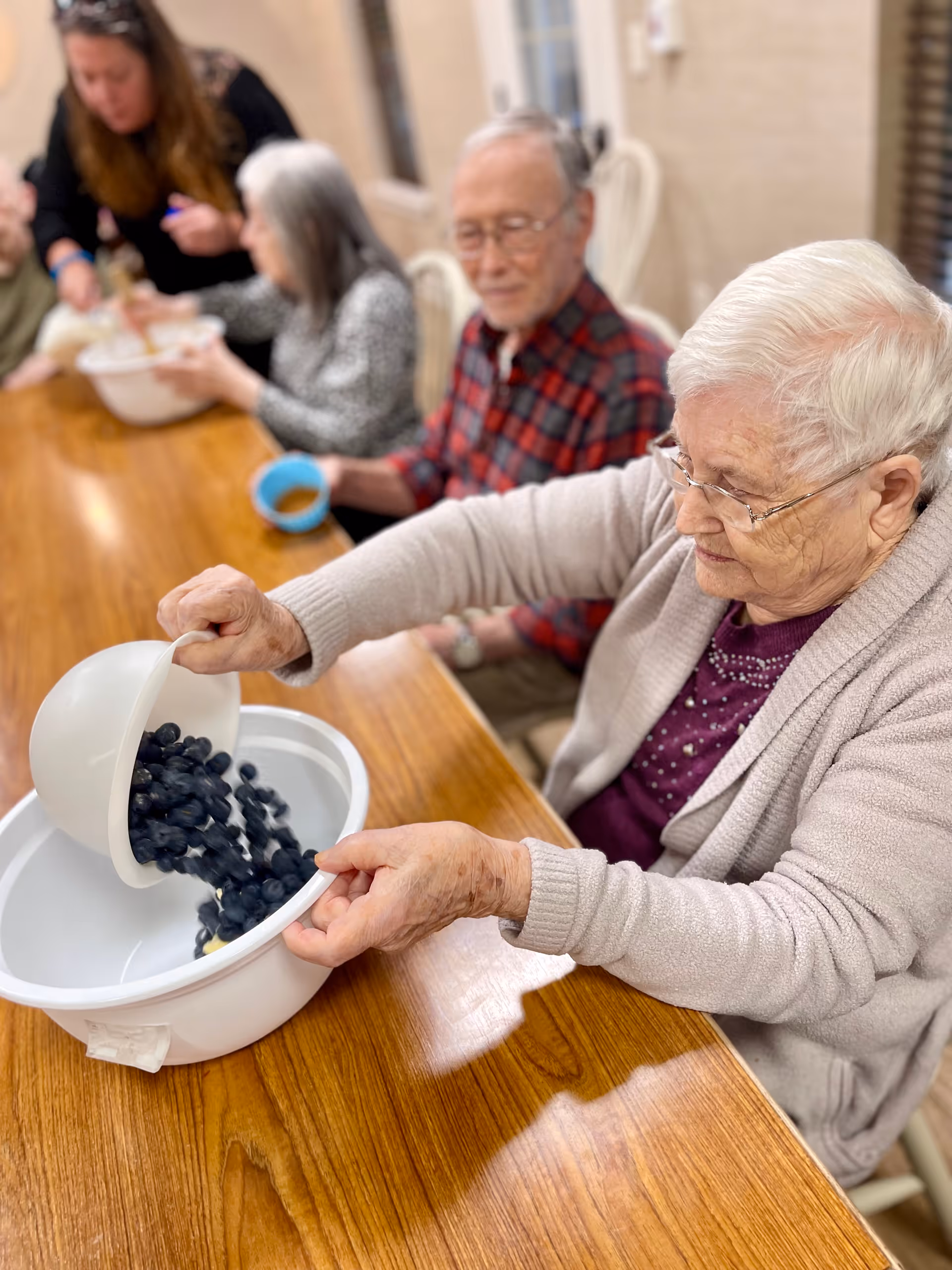 An elderly woman pours blueberries from a bowl into a larger mixing bowl while other seniors sit at a wooden table in a communal dining area.