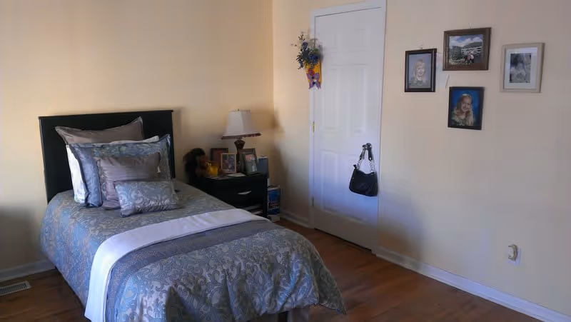 A neatly made bedroom with a patterned blue comforter, nightstand, lamp, and wall photos.