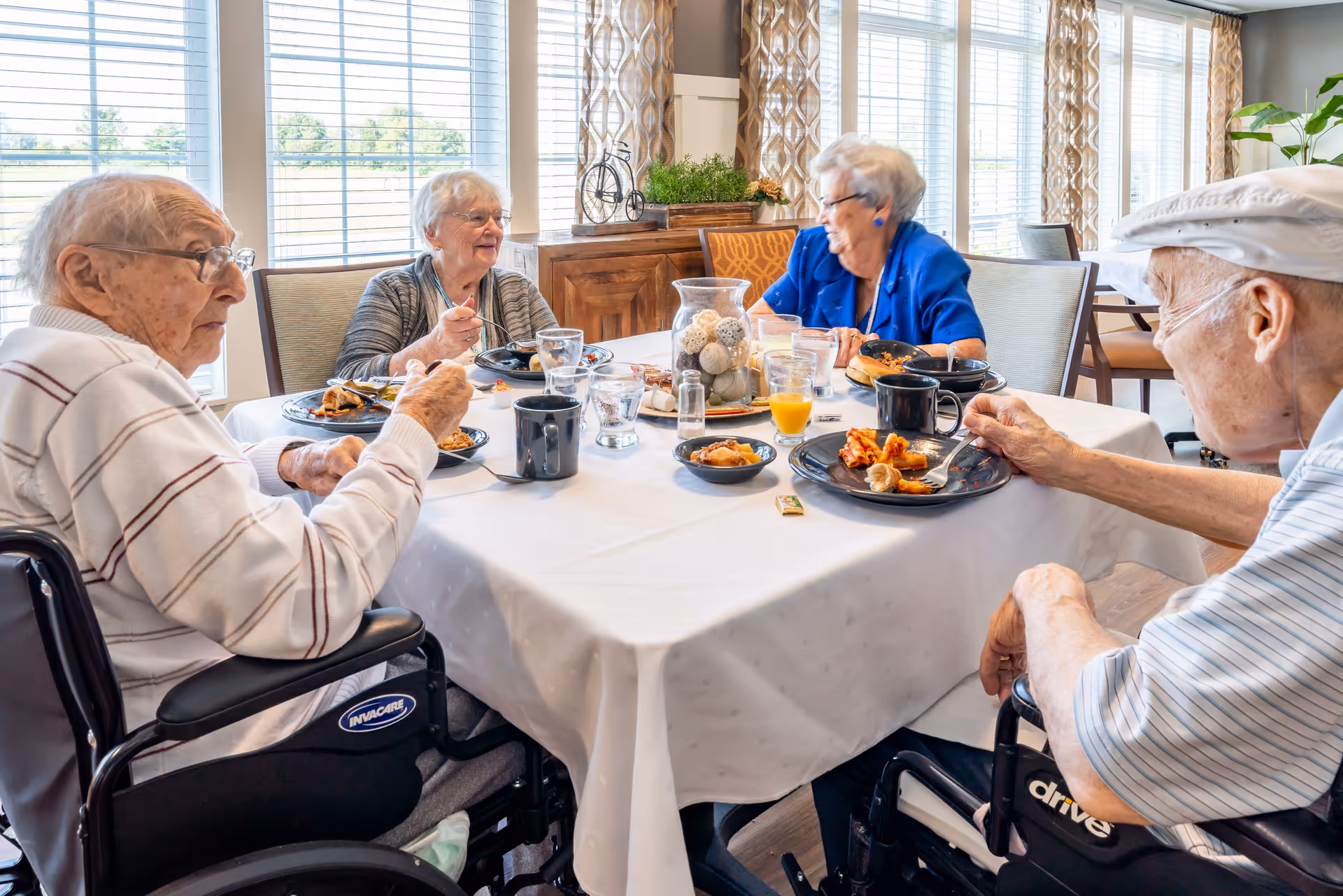 Four elderly individuals sitting around a dining table in a well-lit room with large windows, enjoying a meal together. The table is covered with a white tablecloth and has plates of food, glasses of water, coffee mugs, and a centerpiece with decorative items.