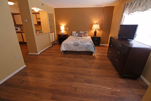 A bedroom with a bed centered against a brown accent wall, flanked by two nightstands each with a lamp. To the right is a dark wooden dresser with a TV on top, positioned next to a window with a valance. The floor is wood, and an open doorway on the left leads to a kitchen area.