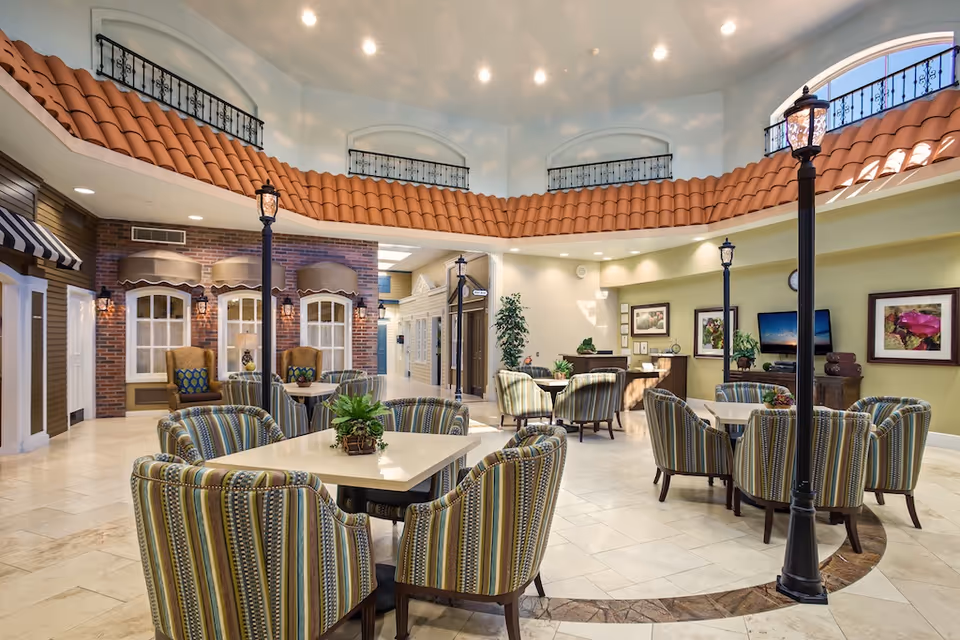 Bright indoor common area with multiple tables surrounded by striped upholstered chairs, decorative streetlamps, and a faux tiled balcony overhead.