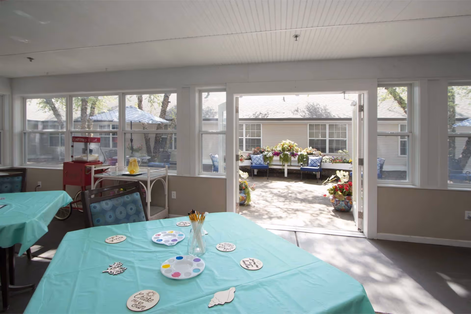 A bright indoor room with tables covered in turquoise tablecloths, some with paint palettes, brushes, and wooden craft pieces. Large windows and an open door lead to an outdoor patio area with potted plants and blue cushioned chairs.