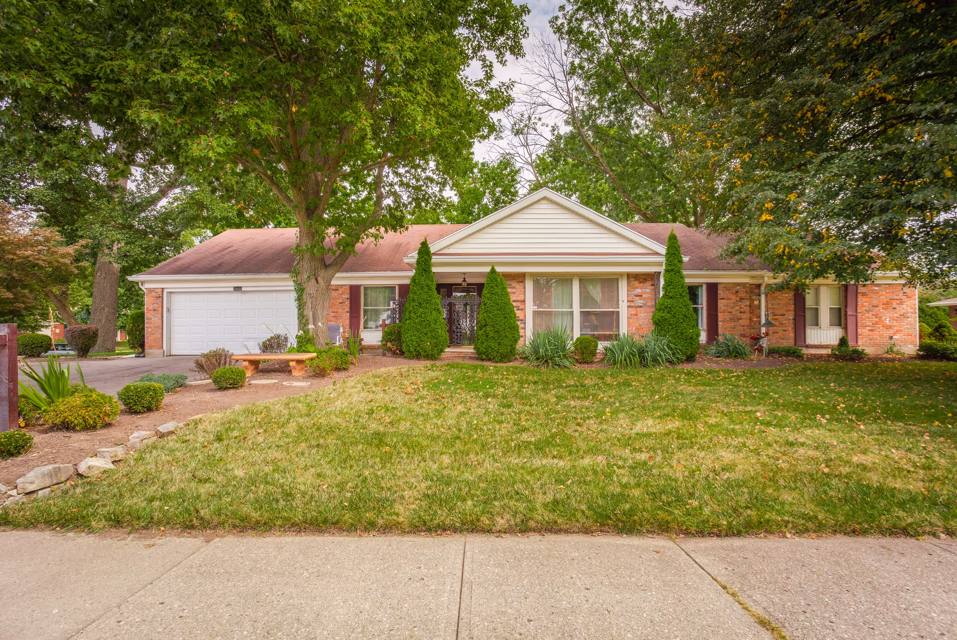 Front exterior view of a single-story brick house with a white garage door, a large tree in the front yard, trimmed bushes, and a concrete sidewalk in front.