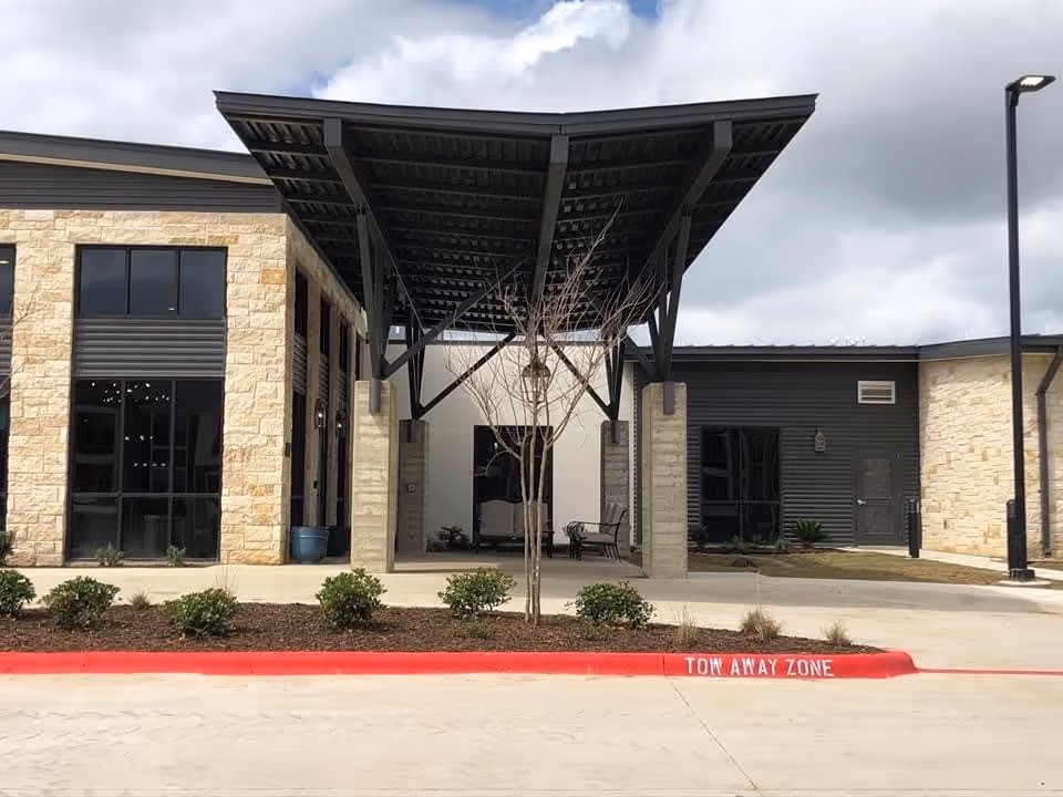 Exterior view of a building entrance with a large covered canopy supported by metal beams and stone pillars. The building features stone and dark siding walls, large windows, a small landscaped area with bushes, and a red curb marked 'TOW AWAY ZONE'. The sky is partly cloudy.