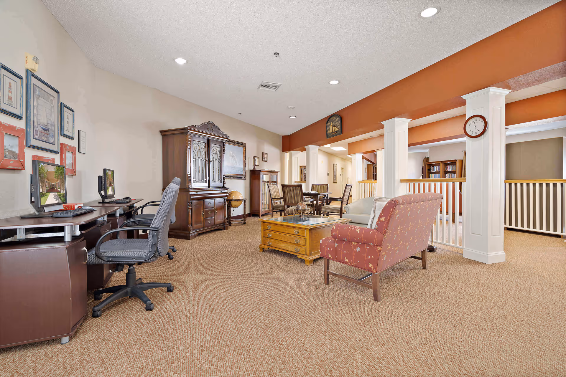 A spacious common area in a senior living facility featuring a carpeted floor, two computer desks with chairs on the left, a wooden cabinet with glass doors, a globe, a coffee table with a glass top, a red patterned armchair, and a small dining table with four chairs in the background. The walls are painted beige with an orange accent beam and white columns, and a clock is mounted on one of the columns.