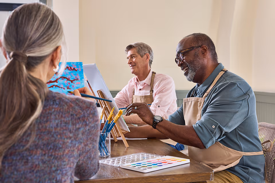 Three seniors engaged in a painting activity at a table indoors. Two men and one woman are seated, each with an easel and paintbrushes, smiling and enjoying the creative session. The setting appears bright and welcoming.