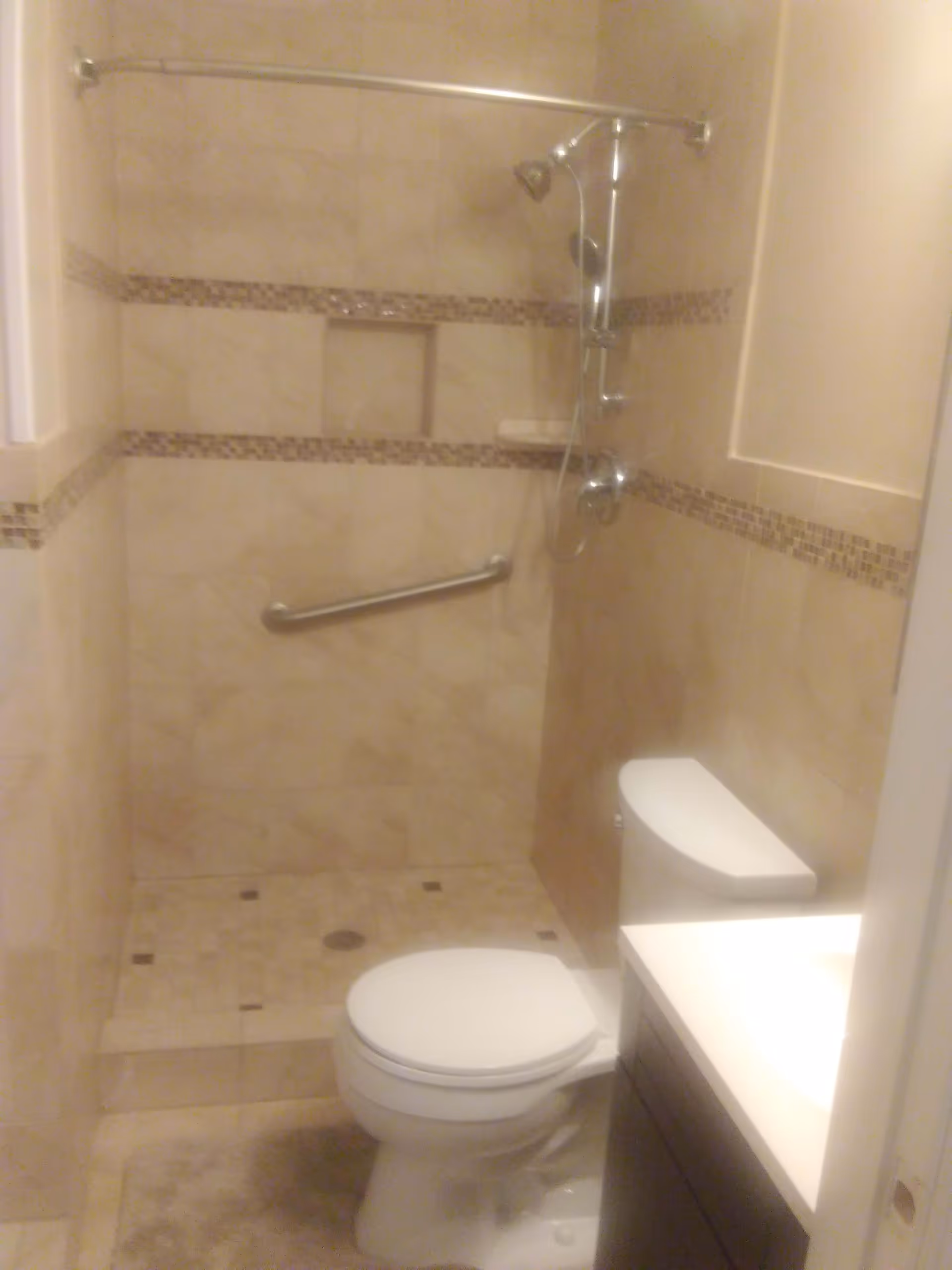 A small bathroom with beige tiled walls and floor, featuring a white toilet, a vanity with a white countertop, and a walk-in shower with a grab bar and a showerhead mounted on the wall.