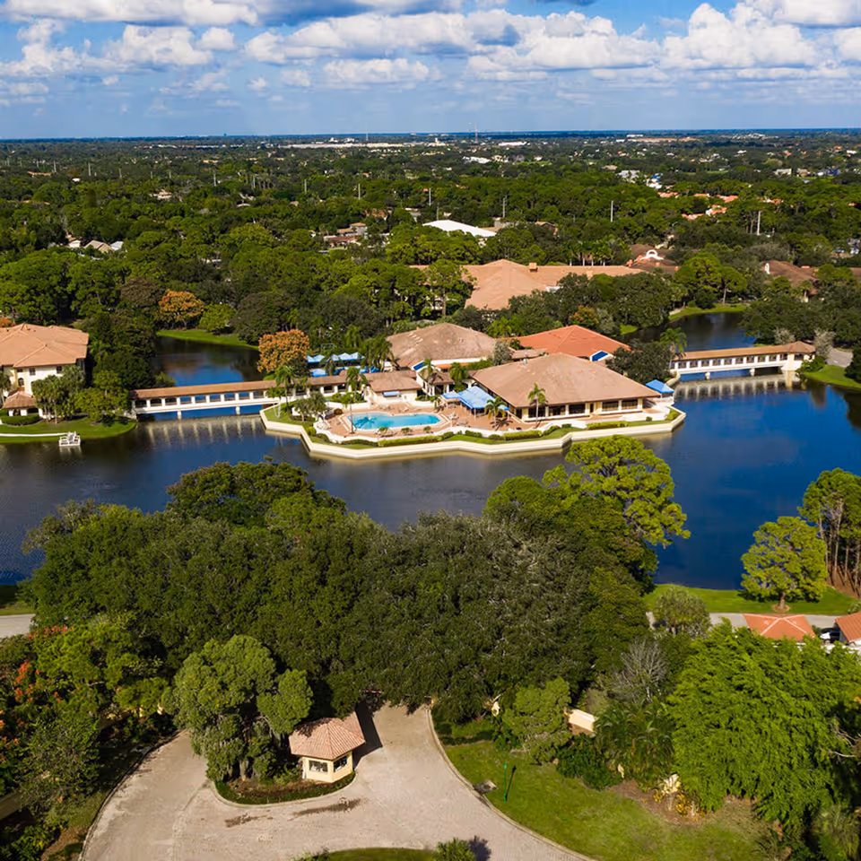 Aerial view of a lakeside senior living community with tan-roof buildings, a central pool on a peninsula, walkways and surrounding trees.